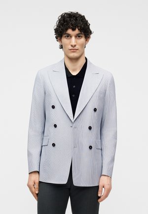 Tânăr cu păr creț, purtând un sacou dublu cu nasturi gri deschis peste o cămașă neagră cu guler, stând în fața unui fundal simplu.