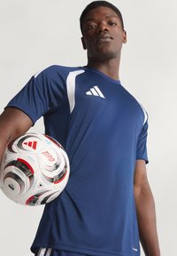 Μπλε ναυτικό αθλητικό μπλουζάκι με κοντό μανίκι και λευκές λεπτομέρειες με το λογότυπο της Adidas. Μια κλασική ποδοσφαιρική μπάλα κρατιέται στο αριστερό χέρι του μοντέλου.