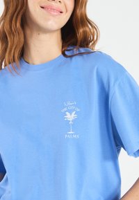 Light blue cotton t-shirt featuring a white graphic print of a palm tree with the text "Welcome to The City of PALMS" beneath. Short sleeves.
