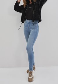 Kvinna i ljusblå skinny jeans, svart jacka, svart tröja med vit text och beige sneakers, som står framför en enfärgad ljus bakgrund.