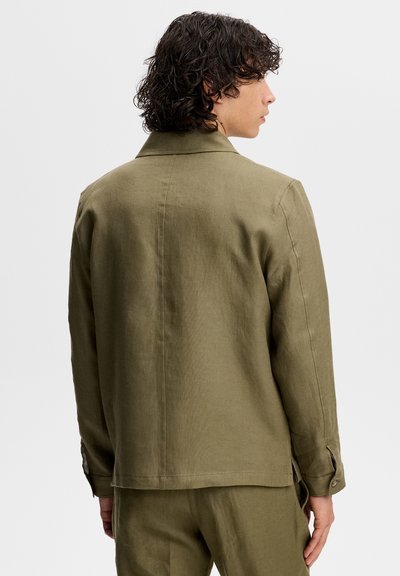 Osoba o kręconych ciemnych włosach, ubrana w oliwkowozieloną koszulę z długim rękawem i dopasowane spodnie, stojąca bokiem na jednolitym tle.