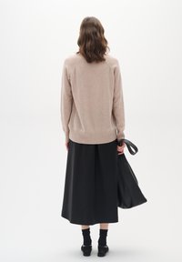 Beige Strickpullover mit geripptem Saum, kombiniert mit einem schwarzen, gefältelten Rock. Eine Textiltasche hängt von einer Hand und zeigt eine glatte Lederstruktur.