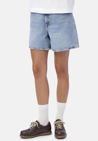 Persona che indossa pantaloncini di denim azzurro chiaro, calzini bianchi alti e sneaker in pelle marrone scuro con lacci chiari su sfondo bianco.