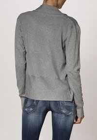 Cárdigan gris con canalé de diseño ajustado, que presenta un fondo de banda y mangas largas, combinado con jeans de mezclilla azul oscuro.