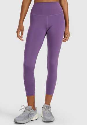 Kobieta nosząca krótkie legginsy w kolorze fioletowym z wysokim stanem oraz szare sportowe buty, stojąca na jednolitym tle.