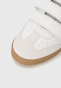 White sneaker featuring a smooth leather upper with suede accents, Velcro straps, and a tan rubber sole, complemented by perforated details.