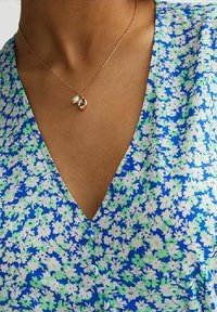 Květinový top s výstřihem do V s modrým, zeleným a bílým vzorem. Je vyroben z lehkého materiálu s hladkou texturou. Jemný zlatý náhrdelník s přívěsky.