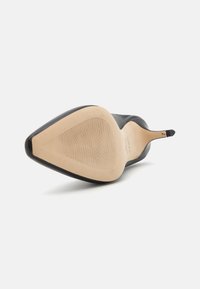 Zwarte hoge hak schoen met een spitse neus, een glad leren bovenzijde en een beige gestructureerde zool met ribbels voor grip.