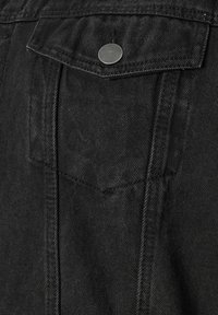 Schwarze Denimjackentasche mit Klappe, gesichert durch einen einzigen silbernen Metallknopf und sichtbaren Nahtdetails.