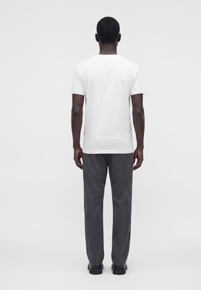 Homme debout, dos à l'appareil photo, portant un t-shirt blanc uni à manches courtes, un pantalon gris foncé et des chaussures noires sur un fond clair uni.