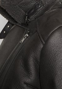 Giubbotto in pelle nera con finitura testurizzata, dotato di colletto in pelliccia sintetica, chiusura a zip, accessori metallici e dettagli con chiusura a scatto.