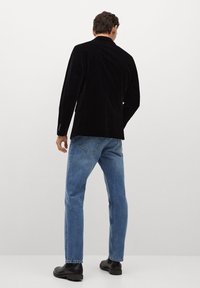 Uomo in giacca nera, jeans blu e scarpe nere in piedi di fronte a un muro bianco con le mani rilassate.