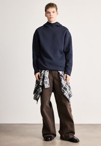 Námořnická mikina s vysokým límcem, ve spojení s volnými hnědými kalhotami. Černobílá kostkovaná košile uvázaná kolem pasu. Černé boty.