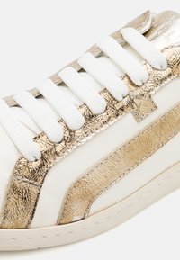 Baskets blanches avec des accents métalliques dorés, présentant une finition texturée, des lacets plats blancs et un design à bout rond.