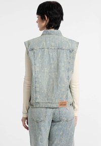 Jasnoniebieska kamizelka dżinsowa o wyblakłej fakturze, bez rękawów, z metką na plecach. W zestawie z pasującymi jasnoniebieskimi spodniami dżinsowymi.