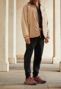 Chaqueta bomber beige con cuello negro, camisa negra de botones, pantalones negros ajustados y zapatillas deportivas multicolores con detalles de malla.
