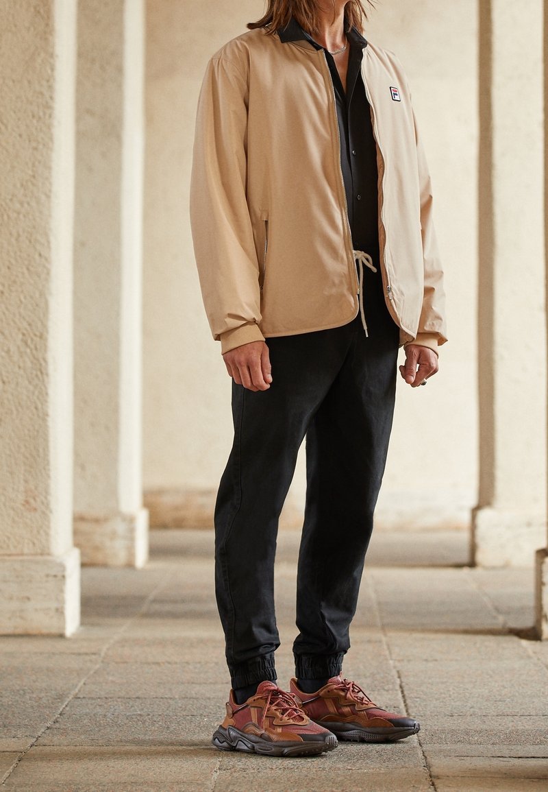 Chaqueta bomber beige con cuello negro, camisa negra de botones, pantalones negros ajustados y zapatillas deportivas multicolores con detalles de malla.