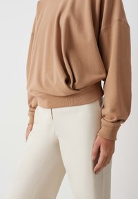 Felpa oversize marrone con polsini e orlo a costine, tessuto texturizzato, abbinata a pantaloni a gamba larga di colore chiaro.
