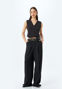 Haut sans manches noir à encolure en V avec cinq boutons, associé à un pantalon large noir taille haute et une ceinture assortie.