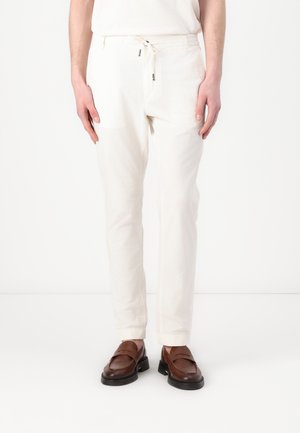 Pantaloni leggeri bianchi con vita regolabile con coulisse, tasche laterali e vestibilità rilassata, abbinati a mocassini marroni su uno sfondo neutro.