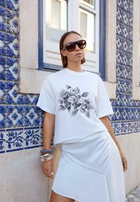 Kvinde iført store mørke solbriller, hvid t-shirt med sort rosetryk, hvid nederdel og armbånd, der læner sig op ad en blåflisevæg under et vindue.