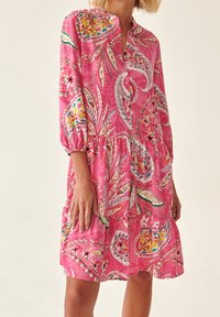 Vrouw in een losse roze jurk met kleurrijke bloeiende en paisleypatronen, driekwartmouwen en een gelaagde rok.