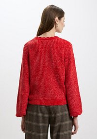 Maglione rosso a maglia con texture a coste, scollatura ondulata e maniche a palloncino. Indossato con pantaloni a quadri, che mostrano un look informale.