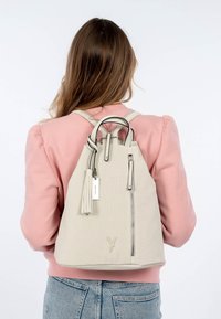 Mochila perforada beige claro con un frente con cremallera, correas ajustables, un acento de borlas y una pequeña etiqueta. Textura suave y forma estructurada.
