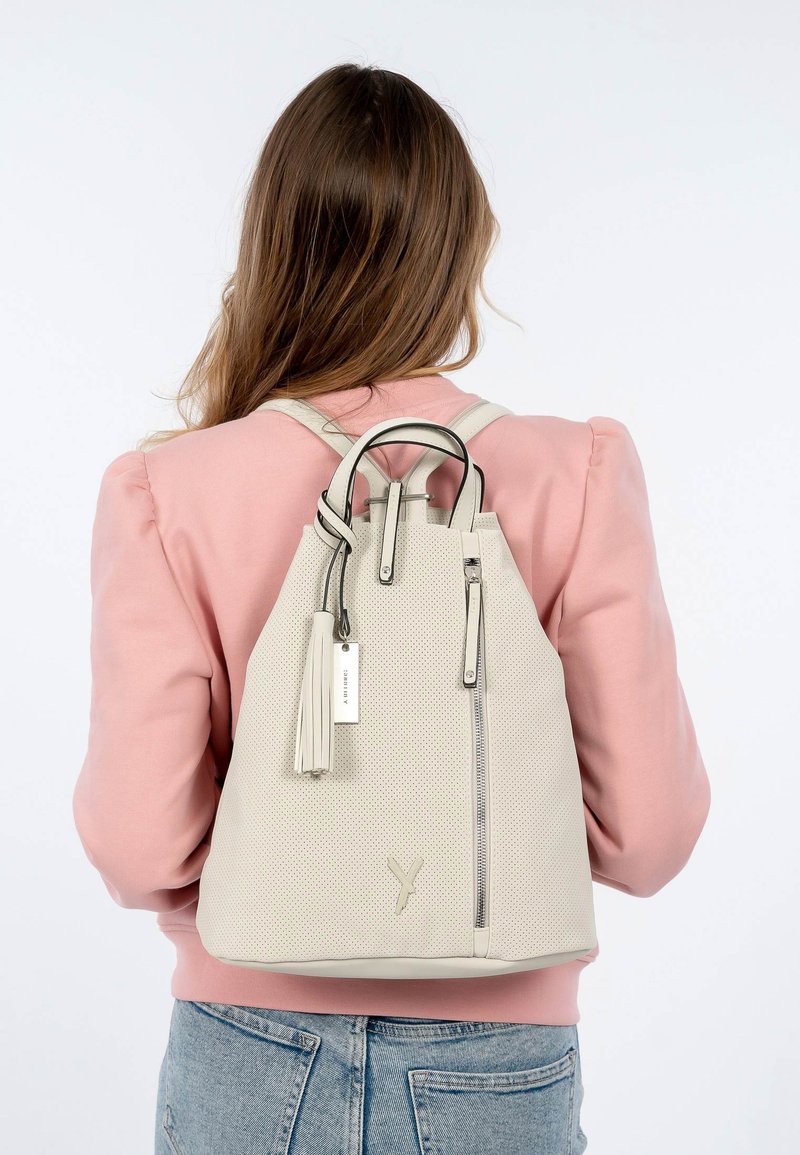 Zaino perforato beige chiaro con una tasca zip anteriore, spallacci regolabili, accentuato da un pendente e un piccolo cartello. Texture morbida e forma strutturata.