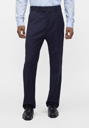 Hombre con pantalones de vestir azul marino, camisa azul claro abotonada y zapatos de vestir de cuero negro, de pie sobre un fondo sencillo.