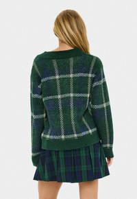 Frau mit langen blonden Haaren, die einen grün-blauen karierten Pullover und einen passenden plissierten Rock trägt, von hinten gegen einen weißen Hintergrund gewandt.