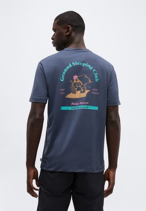 Άνδρας που φοράει μπλε μπλούζα με πολύχρωμο γραφικό και κείμενο «Ground Sleeping Club», προωθώντας το να μένει δροσερός και να κοιμάται έξω.