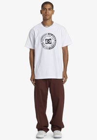 Biely bavlnený tričko s kruhovou čiernou grafikou a textom "DC Shoe Company", v kombinácii s voľnými hnedými nohavicami a teniskami.