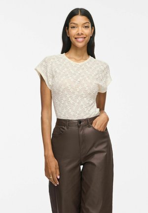 Femme aux longs cheveux noirs portant un haut blanc en dentelle à manches courtes et un pantalon marron taille haute, souriante, une main dans la poche.