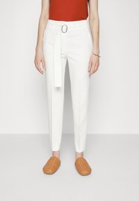 Witte op maat gemaakte trousers met een riem en een zilveren, ronde gesp, gedragen met een roestkleurige top en tan kleurige, geweven instappers.