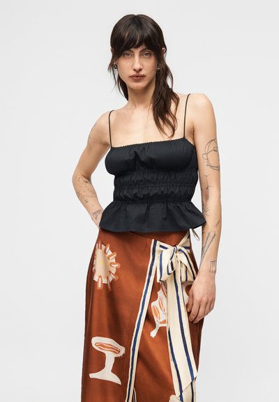 Γυναίκα με τατουάζ που φοράει μαύρο μανίκωτο τοπ με ρυτίδες και καφέ φούστα τύπου wrap με κρεμ λουλουδένιες λεπτομέρειες και φιόγκο, στέκεται μπροστά σε απλό φόντο.