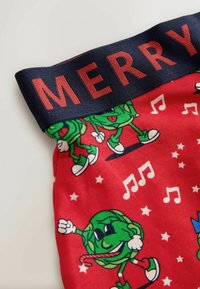 Červené látkové šortky s veselým dizajnom, na ktorom sú zelené postavy, hudobné noty a logo "MERRY" na námorníckom opasku.