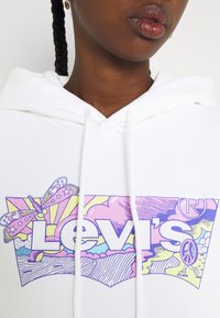 Primer plano de una sudadera blanca con un colorido logo de Levi's que presenta una mariposa, rayos de sol, nubes y un personaje con un signo de paz caminando.