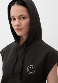 Czarna, bezrękawnikowa bluza z kapturem z okrągłym białym logo przedstawiającym uśmiechniętą buźkę i tekst, prezentująca miękką tkaninę i luźny krój.