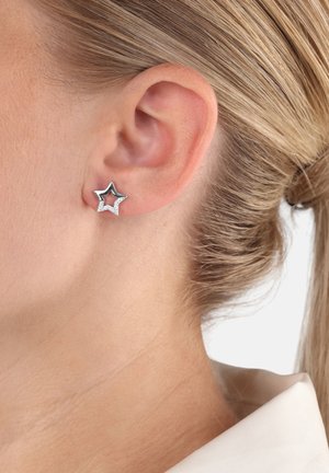 Pendiente de estrella plateada en forma de Stud, llevado en la oreja izquierda de una persona con el cabello rubio recogido, sobre un fondo blanco.
