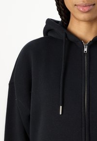 Sweatshirt preto com fecho zip, confeccionado em tecido suave, com cordões ajustáveis e fecho de metal prateado. Ajustado ao corpo, com um design simples.