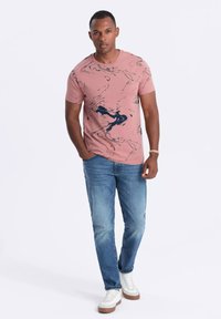 T-shirt rosa a maniche corte con un motivo astratto blu navy, abbinato a jeans azzurri chiari e sneakers bianche con dettagli marroni.