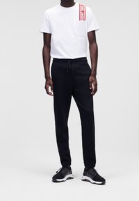 Vit bomullströja med en röd logotryck och ficka, kombinerad med svarta joggingsbyxor med elastisk midja och sidfickor, i sällskap med svarta sneakers.