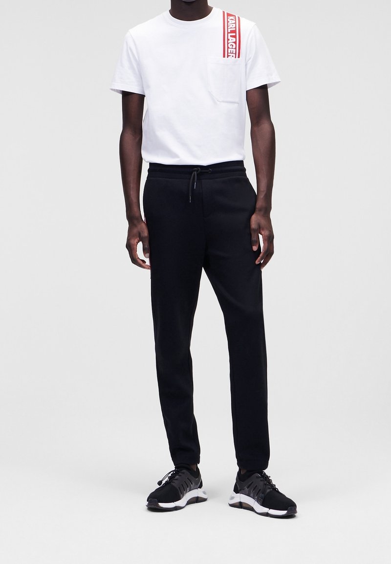 Vit bomullströja med en röd logotryck och ficka, kombinerad med svarta joggingsbyxor med elastisk midja och sidfickor, i sällskap med svarta sneakers.