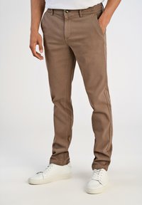 Uomo che indossa pantaloni slim-fit marroni con tasche laterali e posteriori, abbinati a sneakers bianche con lacci, in piedi su uno sfondo semplice.