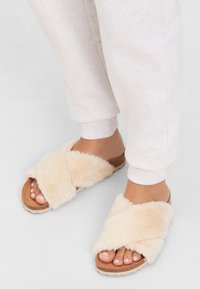 Beige cross-strap slippers met een pluche, zachte textuur en een kurken zool. Blootgestelde tenen tonen een natuurlijke nagelkleur. Gezellig huisfootwear.