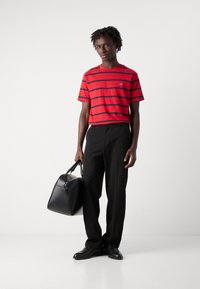 Rød og marineblå stribet kortærmet t-shirt, sorte skræddersyede bukser, holder en sort læder sportstaske, stående på en hvid baggrund.