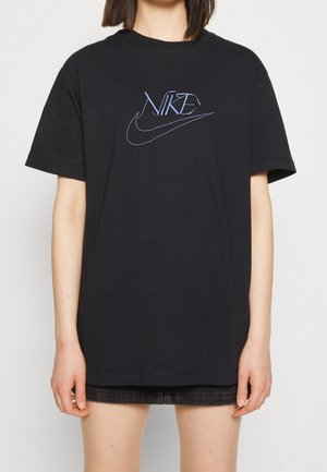 Osoba nosząca czarną, oversize koszulkę Nike z niebieskim haftowanym logo oraz czarne szorty, stojąca na jednolitym jasnym tle.