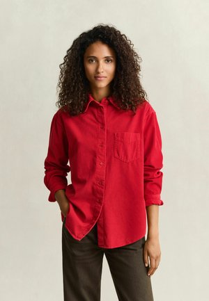 Femme aux cheveux bouclés portant une chemise rouge vif à boutons avec les manches retroussées et un pantalon sombre, debout devant un fond clair uni.