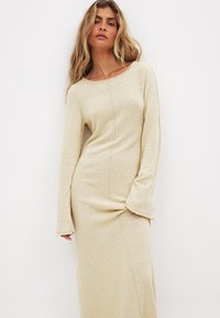 Robe en tricot beige longue avec encolure ronde, manches longues et silhouette ajustée. Présente des coutures visibles et une texture douce.